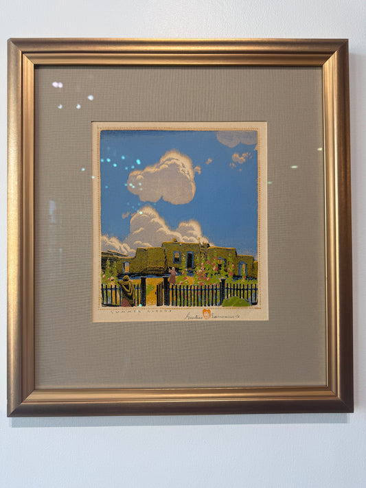 A framed and signed limited edition print by celebrated artist Gustauve Baumann featuring a colorful illustration of a green adobe-style building under a bright blue sky with white clouds, mounted in a gold-toned frame with a beige mat on a light wall.