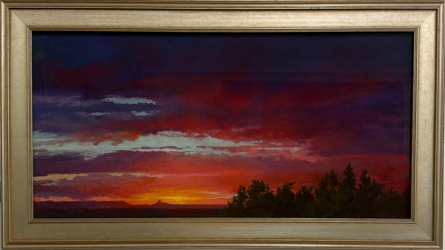 A framed landscape oil painting depicting a dramatic sunset or sunrise with dark blue, red, and orange clouds above a dark tree line, housed in a wide, textured gold frame.