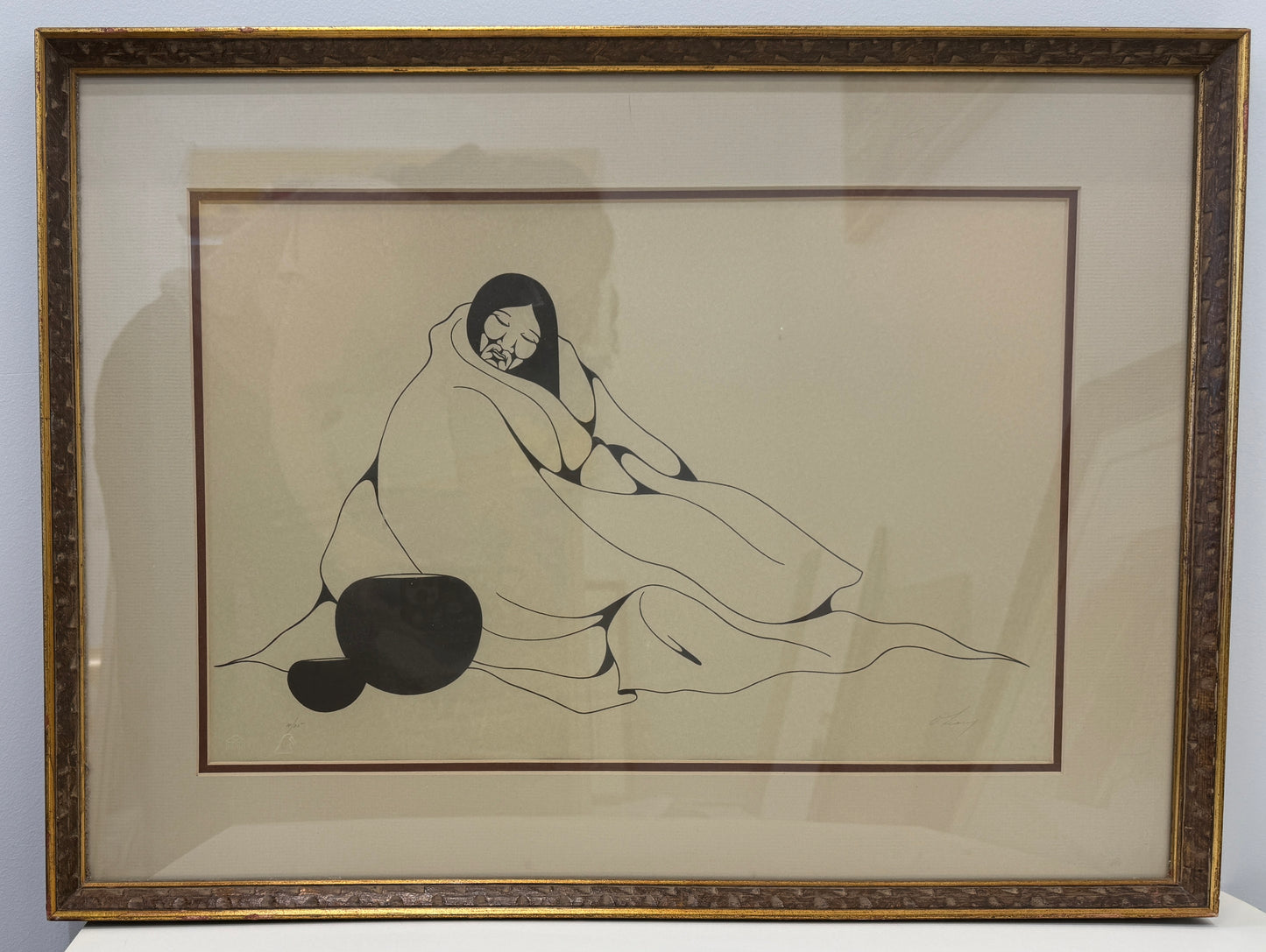 A framed black-and-white drawing titled "Indian Woman 10/25" depicts a person sitting wrapped in a blanket, with two round objects nearby against a plain background. The artwork’s framed dimensions are 20x27.